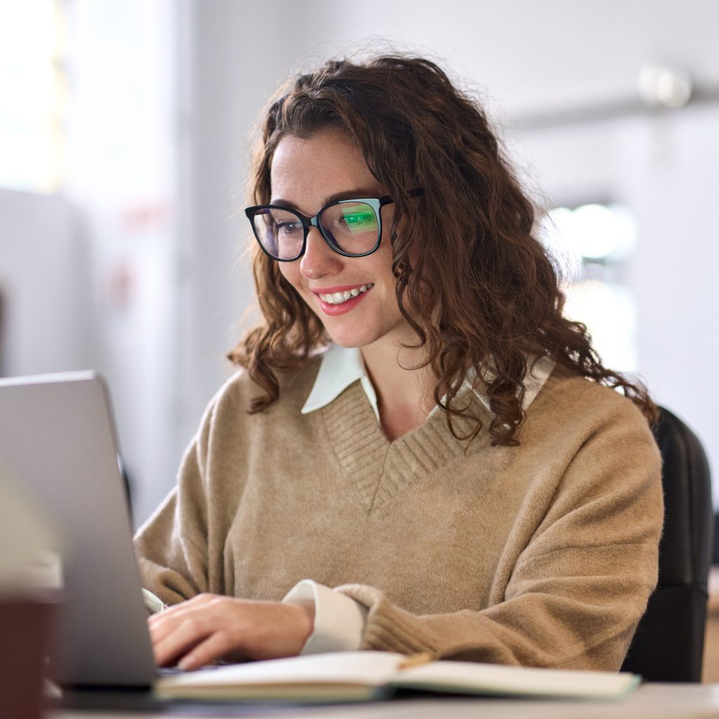 Person seated with comfort at a desk using a laptop computer, with an open notebook on the desk and a softly lit indoor workspace in the background.