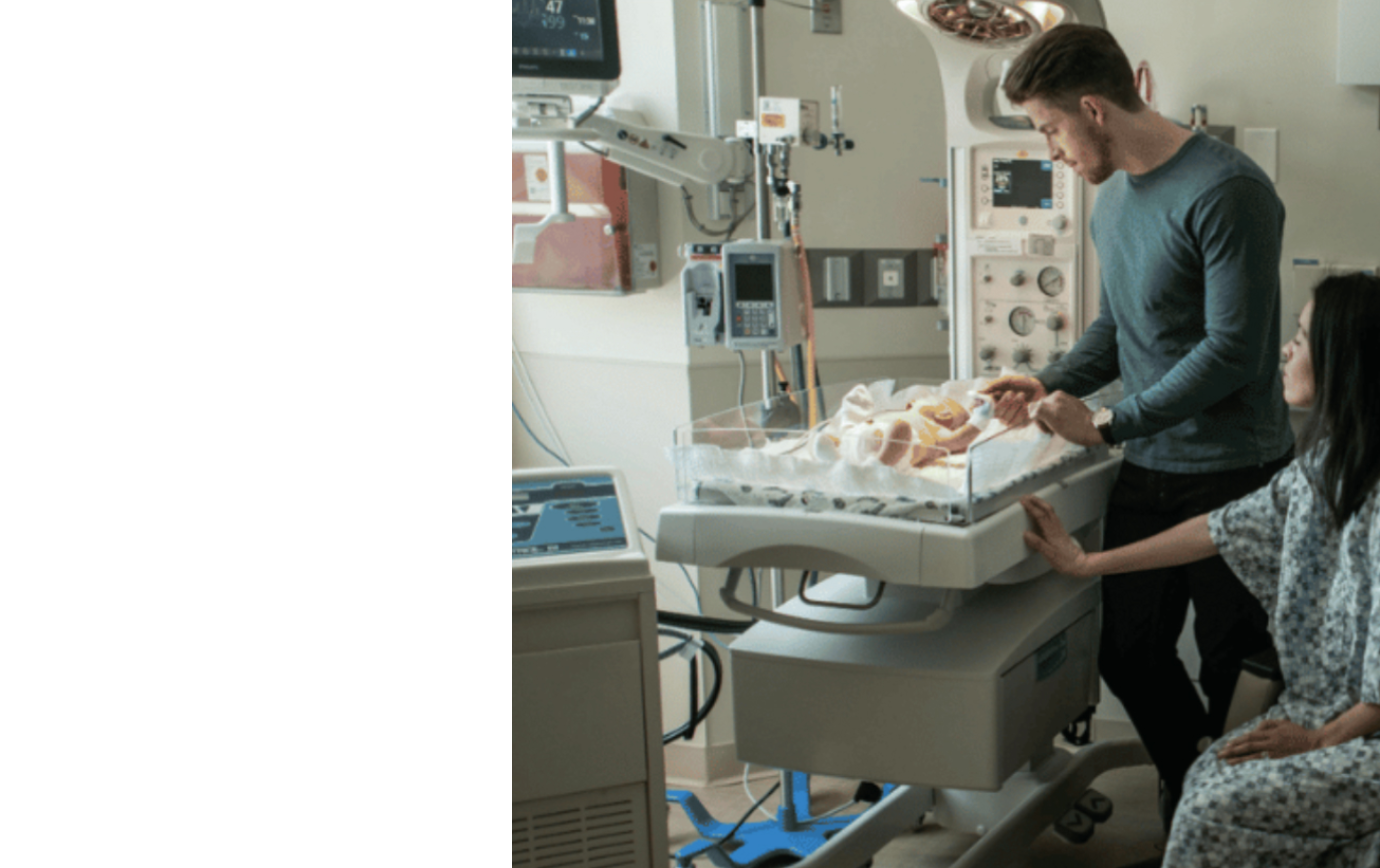 Baby placed on Gentherm cooling blanket with Blanketrol device. Mom and Dad standing beside the bassinet.